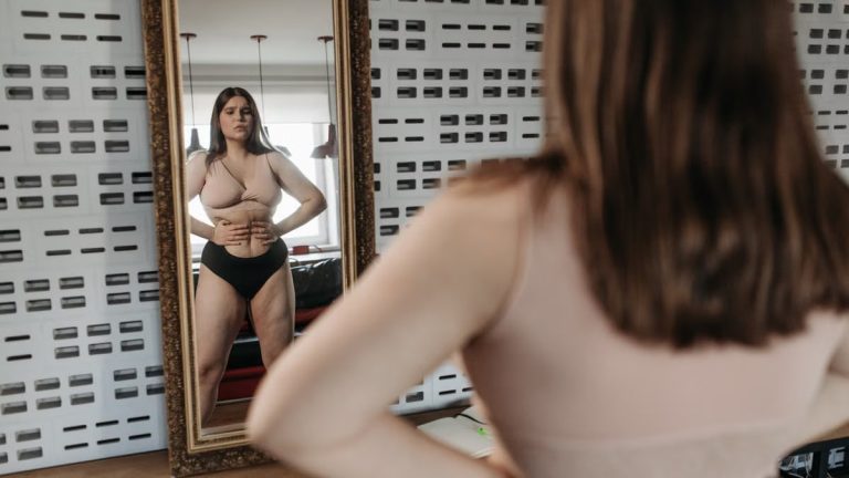 Woman in underwear looking at her reflection in a mirror, contemplating body image.