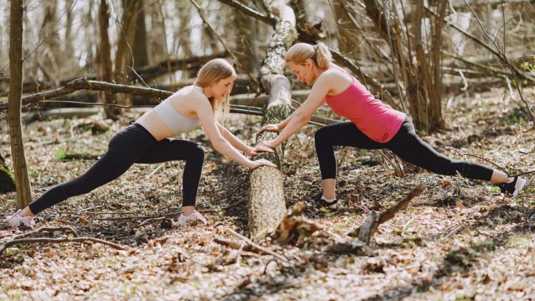 Side view of young female athletes in activewear stretching legs together using fallen tree during fitness workout in spring forest adhering to healthy lifestyle